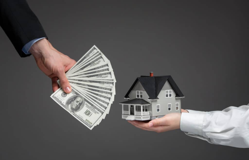 Hand Holding A Wad Of Hundred Dollar Bills Reaching Out To Another Hand Holding A Model Of A House