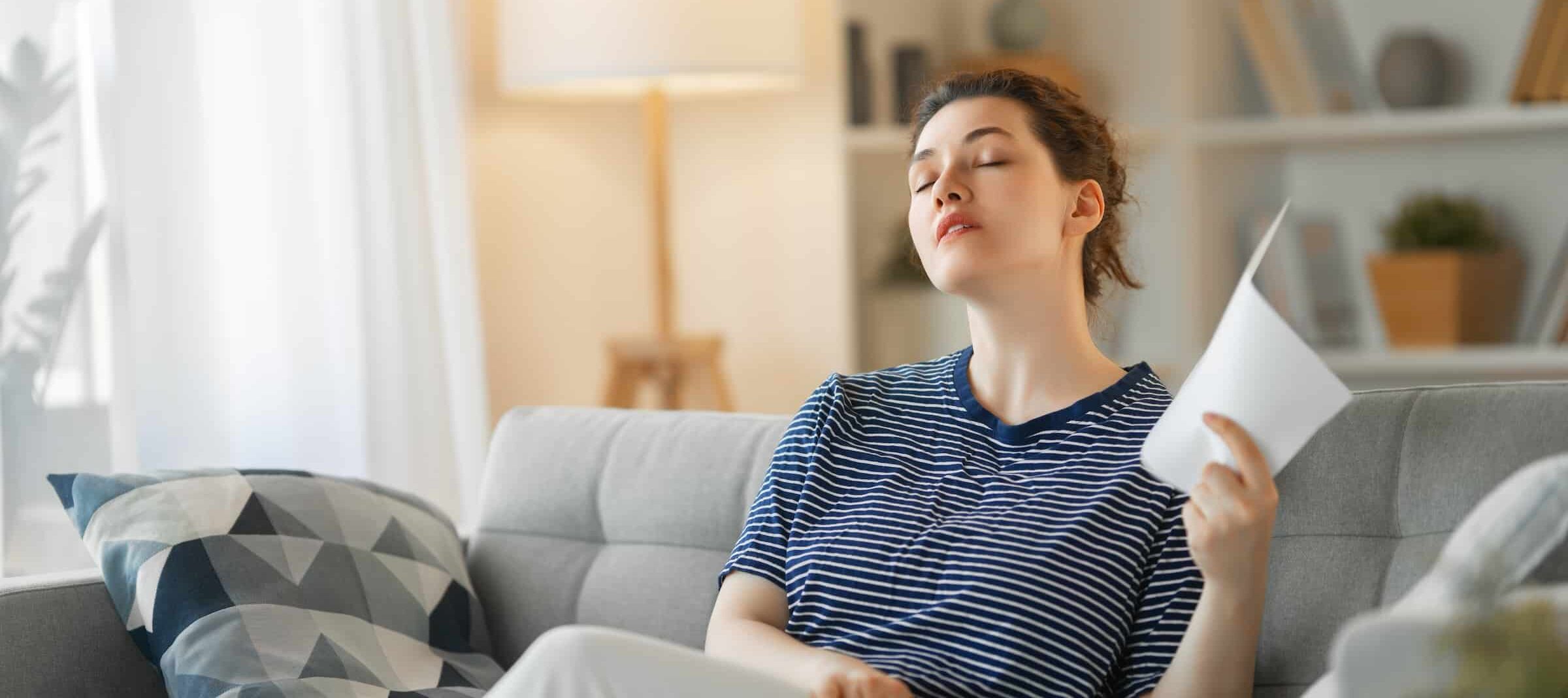 Uncomfortable looking woman, fanning her face with a piece of paper while sitting on her couch